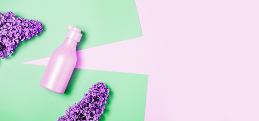 Creative minimal beauty and health background with pink bottle and lilac flowers. Minimal top view and flat lay of cosmetic on pink and green trendy background.