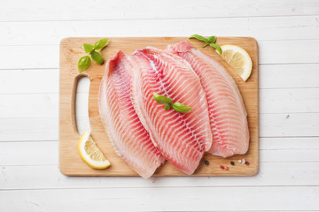 Raw fish fillet of tilapia on a cutting Board with lemon and spices. White table with copy space. - obrazy, fototapety, plakaty
