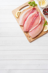 Raw fish fillet of tilapia on a cutting Board with lemon and spices. White table with copy space.