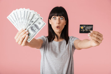 Excited happy woman posing isolated over light pink background wall holding money and credit card.