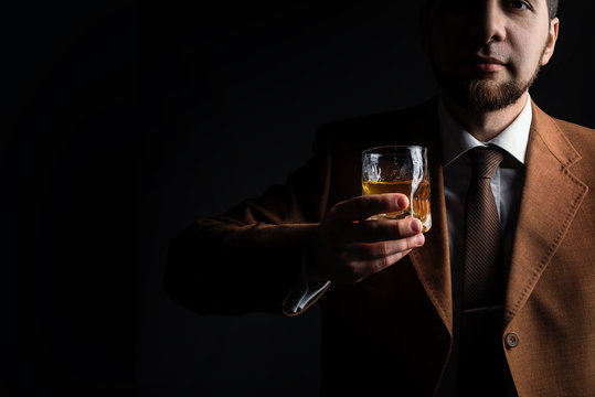 Stylish bearded man in elegant jacket holds whiskey glass and toast in the dark