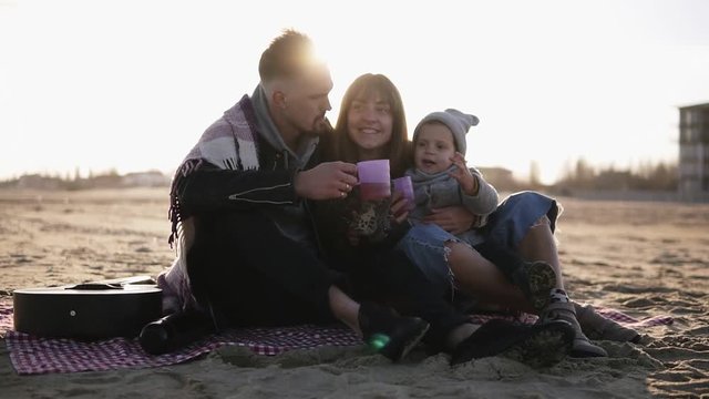 Portrait of young stylish family in casualwear sitting on plaid by waterside and having hot drinks at picnic. Cold weather, weekend recreation. Lens flares on the background