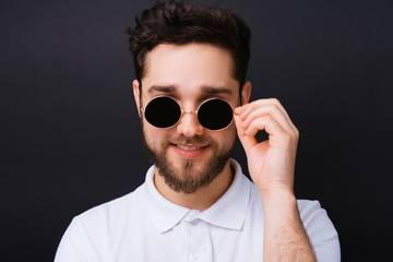 Obraz premium Young attractive bearded man wearing white polo, black vintage, round sunglasses o black background looking through the glasses to the camera.