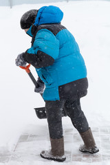 Fototapeta na wymiar Old woman in warm blue jacket clears a snowdrifts with a snow shovel