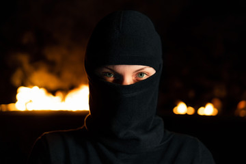 Portrait of female protesting activist in black balaclava against burning barricades at night. Concept of strikes, political conflicts and confrontation