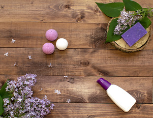 Cosmetic flat lay with a bouquet flowers lilac. Spa set of bottle cream, handmade raging bombs and handmade lavender soap on wooden slice on brown wooden background.