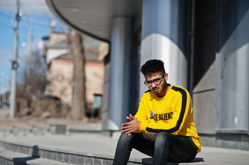 Asian man in eyewear and yellow sweatshirt sitting on stairs against new modern building.