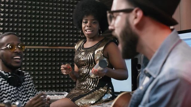 Medium shot of black male sound engineer in glasses clapping his hands while listening to African-American woman with shaker and Caucasian man with guitar performing song in recording studio