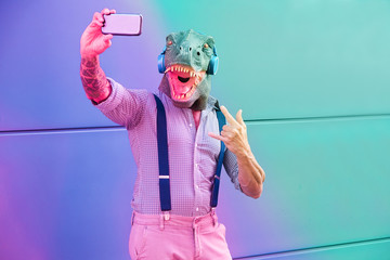 Tattooed man with t-rex mask using smartphone while listening music - Crazy senior male taking...