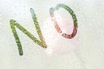 A finger drawing of the shape of the strips of the word no on a semitransparent misted glass. raindrops of spring rain on the window close-up. Blurred background, copy space