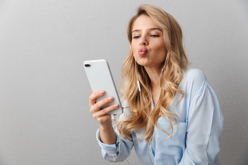 Naklejka premium Young pretty blonde business woman posing isolated grey wall background using mobile phone listening music with earphones talking take a selfie.