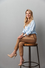 Blonde business woman posing isolated grey wall background sitting on stool.