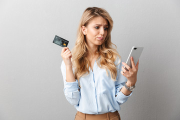 Sad young pretty blonde business woman posing isolated grey wall background holding credit card using mobile phone.