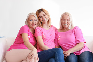Beautiful women of different ages with pink ribbons sitting on sofa in room. Breast cancer concept