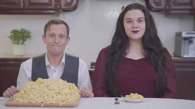 Portrait of a stylish plump woman and modest nerd sitting at a table in front of noodles in the kitchen. Before fat girl there is a small plate and a thin man is a huge plate with pasta. Funny family.