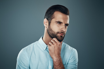 Looking in the future. Handsome young man looking away and keeping hands on chin while standing against grey background