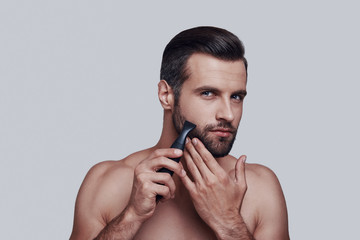 Taking care of his beard. Handsome young man shaving and looking at camera while standing against grey background