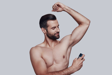 Freshness. Handsome young man applying deodorant and smiling while standing against grey background - obrazy, fototapety, plakaty