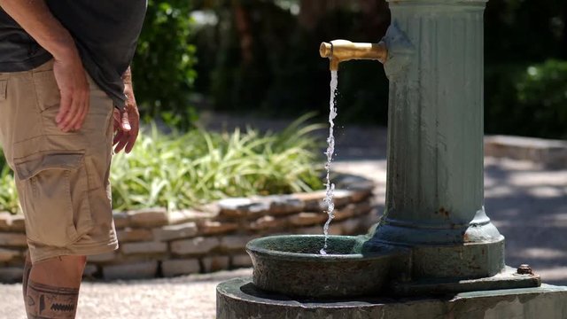 At a historic public tap, a man drinks water and refreshes himself in the palm trees park in the Spanish city of Elche in warm weather and sunshine.