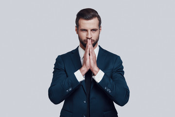 Praying for success. Handsome young man keeping hands clasped while standing against grey background