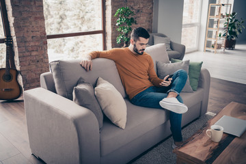 Portrait of his he nice-looking attractive focused concentrated calm bearded guy sitting on divan surfing web addict 5g app dating service at industrial loft brick interior style living-room