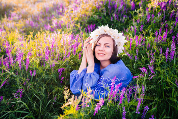 Young pretty woman with flower wreath sitting in purple field. Portrait of girl in blue clothing.