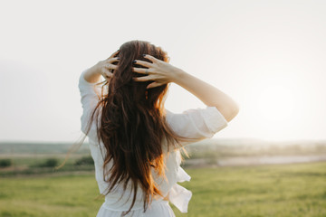 Carefree long hair girl in white clothes on field at sunset, view from back. Sensitivity to nature...