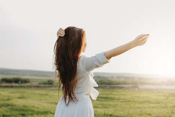Carefree long hair girl in white clothes on field at sunset, view from back. Sensitivity to nature...
