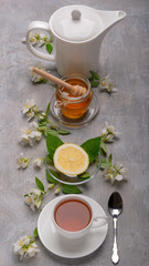 A cup of tea, fresh lemon, a jar of honey, jasmine flowers and a white kettle on a stone background.