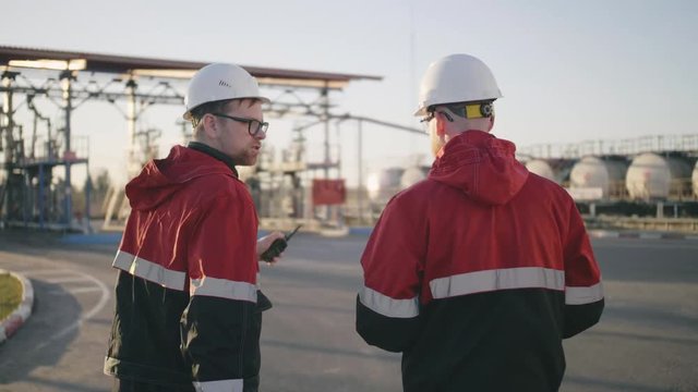 Back side view of oil refinery engineers that walk to fuel tanks, talk and gesticulate