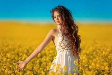 Beautiful carefree girl with long curly healthy hair over Yellow rape field landscape background. Attracive brunette with blowing hairstyle enjoying sunset, outdoor beauty portrait.