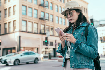 Attractive local woman walking on street and looking at smart phone chatting online conversation. young girl backpacker in sunglasses using cellphone searching direction on internet map app busy city