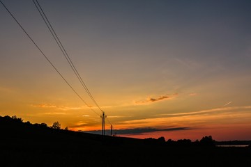 Bright colorful sunset over rural countryside. Spring evening landscape with silhouettes of electric poles and lines. Hilly horizon with trees and blue, orange, pink and yellow sky with clouds. 