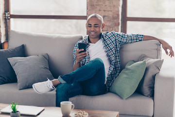 Portrait of positive cheerful trendy stylish guy youth sit divan hold hand device blogger read news dressed denim shirt jeans checked plaid room apartment