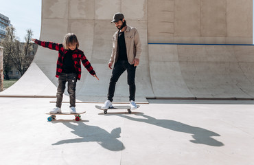 Young father and his son ride skateboards in a skate park with slides outside at the sunny day