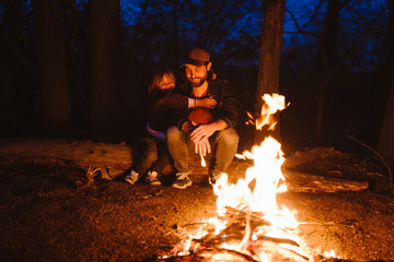 Father and his little son sitting together on the logs in front of a fire at the night. The hike in...