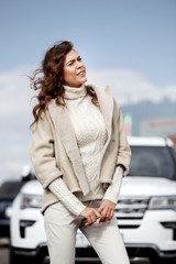 Stylish brunette girl dressed in stylish knitted white sweater, light pants and a short sheepskin coat poses against of a white car in the street