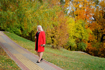 Fototapeta premium An attractive blonde in a red coat and a scarlet rose in her hand poses on a path in the Park on a Sunny autumn day. Dreaming and looking away. Woman's size plus xxl. Horizontal view