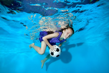 Happy little girl swimming and playing underwater with soccer ball. She smiles and looks at the...