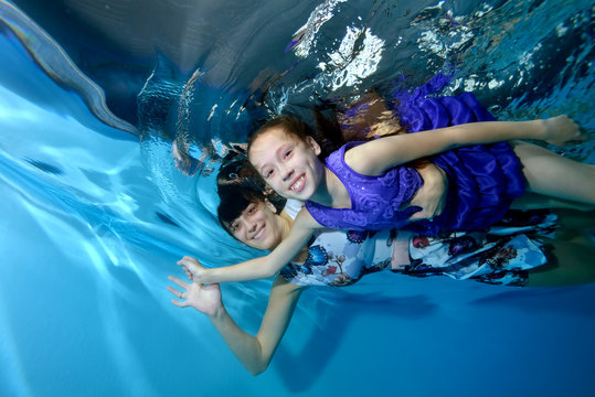 Happy mom and little daughter hugging, floating underwater, holding hands. They smile and look at the camera. Underwater photo on a blue background. Portrait. Horizontal orientation