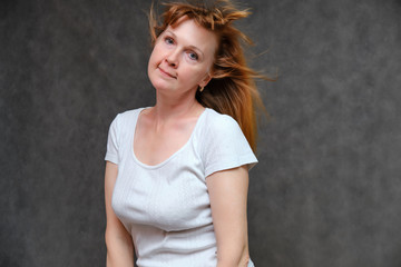 Naklejka premium Studio portrait of a beautiful pretty woman age of 40 years on a gray background, smiling, showing different emotions, happy with life. In a white T-shirt with red hair. Directly opposite the camera.