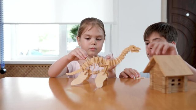 Brother and sister assemble a dinosaur 3d puzzle of wooden parts. The girl gives her brother details and watches with interest the process. Studio video shot with dolly pan of child hobby in 4K