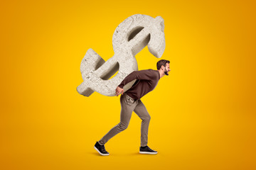 Young man in casual clothes carrying big stone dollar sign on his back on yellow background