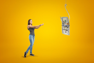 Young brunette girl wearing casual jeans and t-shirt holding out her hands to money dollar on fish hook on yellow background