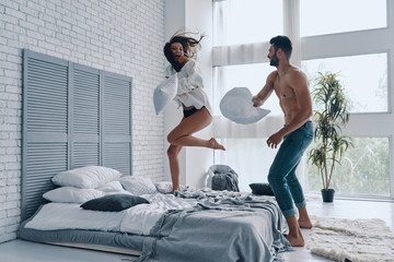 Fun time. Full length of happy young couple having a fun pillow fight while spending carefree time in the bedroom