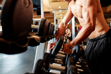 Fototapeta na wymiar Close-up of shirtless bodybuilder taking dumbbell from the shelf and is going to training in gym