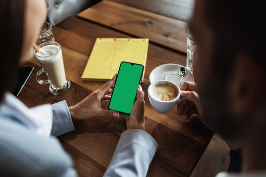 Mockup image of a business people holding smart mobile phone with blank green screen on vintage wooden table in modern cafe restaurant during meeting or lunch.