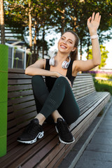 Image of young sportswoman waving hand while sitting on bench with fitness mat in city park