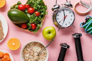 top view of fresh diet food, measuring tape, sport equipment and alarm clock on pink background...