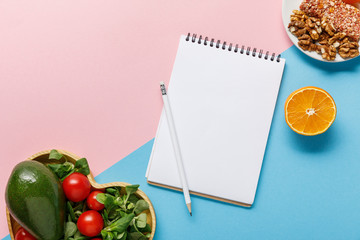 Obraz na płótnie Canvas top view of empty notebook, vegetable salad, orange and nuts on pink and blue background
